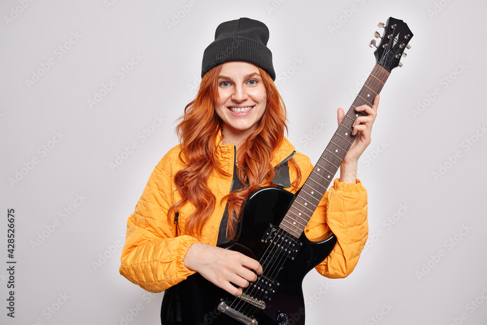 Fototapeta premium Beautiful redhead teenage girl learns to play electric guitar poses with musical instrument smiles pleasantly being pop musician wears trendry hat orange jacket isolated over white background