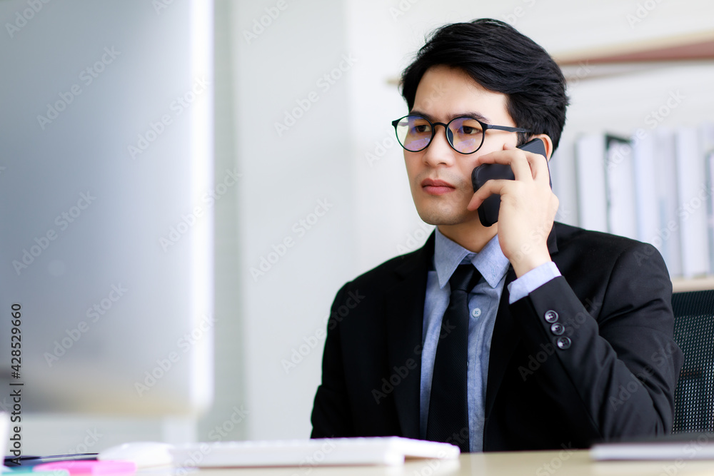 Asian handsome business man wearing eyeglasses, formal black suit with necktie, happily smiling with brace on his teeth while working and talking or communicating on mobile phone in office. Asian handsome business man wearing eyeglasses, formal black suit with necktie, happily smiling with brace on his teeth while working and talking or communicating on mobile phone in office.