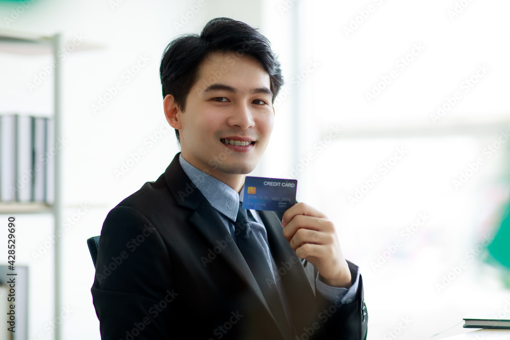 Asian business man wearing formal suit with necktie, happily smiling with brace on his teeth and holding a credit card to present about online purchasing or buying. E-commerce and Financial Concept. Asian business man wearing formal suit with necktie, happily smiling with brace on his teeth and holding a credit card to present about online purchasing or buying. E-commerce and Financial Concept.