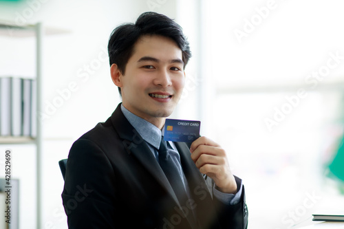 Asian business man wearing formal suit with necktie, happily smiling with brace on his teeth and holding a credit card to present about online purchasing or buying. E-commerce and Financial Concept.