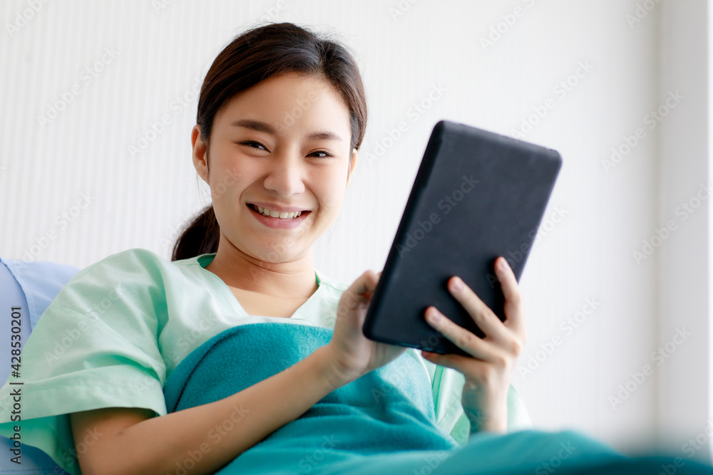 An asian young pretty female patient smiling with happiness and touching screen of tablet for online chat or communicate while lying on bed in hospital. Technology and Medical Concept. An asian young pretty female patient smiling with happiness and touching screen of tablet for online chat or communicate while lying on bed in hospital. Technology and Medical Concept.