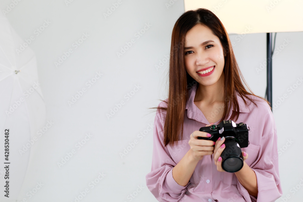 An asian beautiful adult female photographer with long hair wearing casual shirt, holding a camera, posing and standing in photo studio with blur background of  lighting equipments.