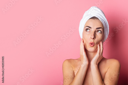 young woman with a towel on her head, bare shoulders, looks up in surprise on a pink background