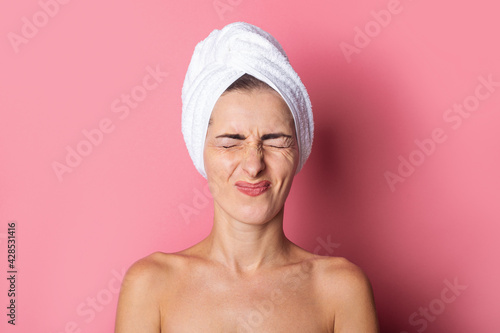 squint young woman with a towel on her head, eyes tightly closed on a pink background