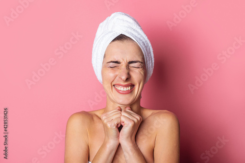 smiling young woman with a towel on her head, eyes tightly closed holds her hands under her chin on a pink background