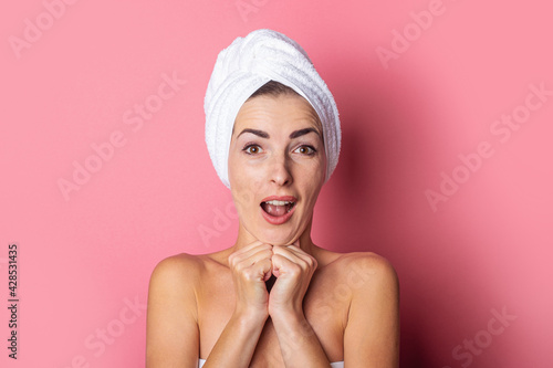 surprised young woman with a towel on her head, bare shoulders, holds her hands under her chin on a pink background