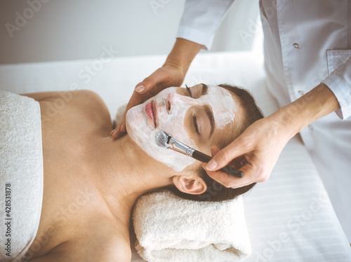 Beautiful brunette woman enjoying applying cosmetic mask with closed eyes comfortable and blissful. Relaxing treatment in medicine and spa center concepts