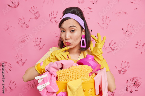Self confident beautiful housewife busy doing cleaning tasks wears headband earrings rubber gloves poses over stack of dirty linen performs regular washing isolated over pink wall with handprints