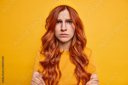 Indoor shot of frustrated ginger teenage girl has gloomy sullen expression feels offended keeps arms folded insulted by cruel words wears hat casual t shirt models against vivid yellow background