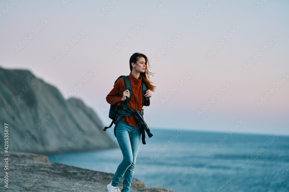 woman in jeans with a backpack on her back sea in the background and high mountains