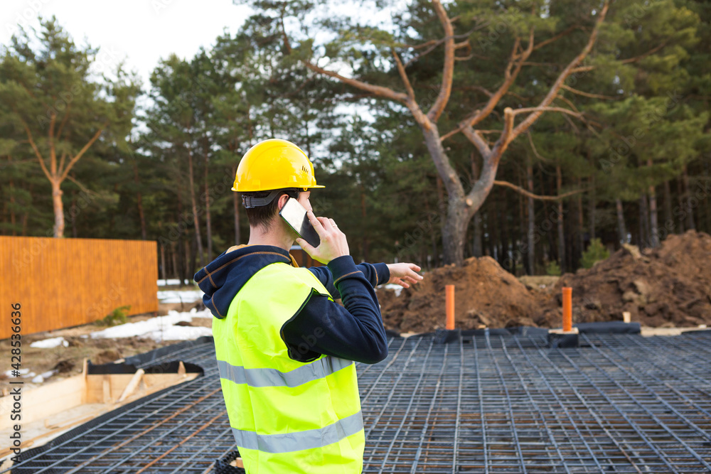 Construction worker talks on a smartphone in a yellow helmet and protective vest against the background of the construction of house-reinforcement for the foundation, pipes and blind area.