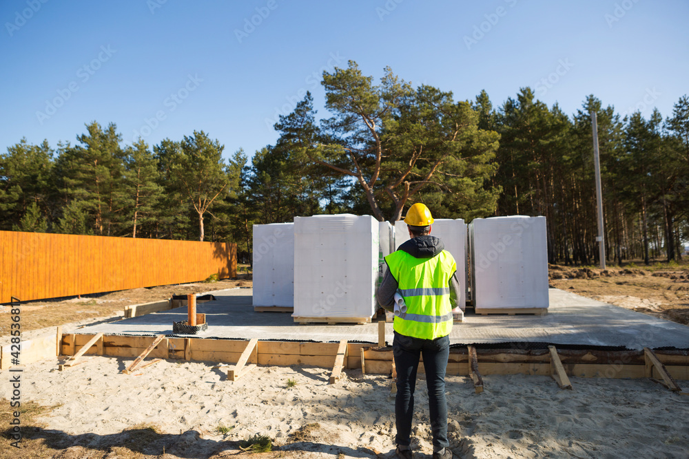 The project architect's back is on the construction site of a house with the foundation laid and the blocks delivered. Construction worker in a protective yellow helmet and a signal vest. Mock up 