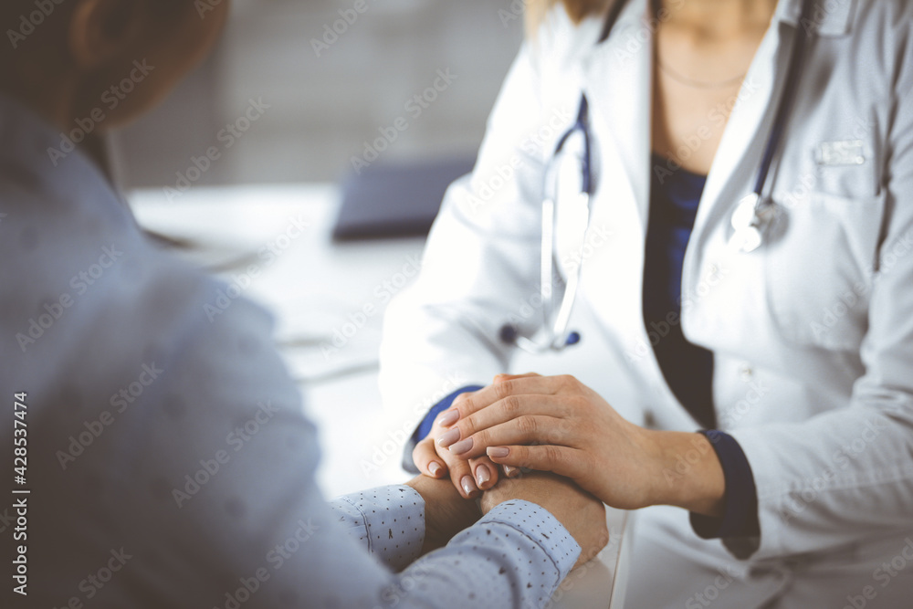 Unknown woman-doctor is holding her patient's hands to reassure the patient, discussing current health examination, while they are sitting together at the desk in the cabinet in a clinic. Female Unknown woman-doctor is holding her patient's hands to reassure the patient, discussing current health examination, while they are sitting together at the desk in the cabinet in a clinic. Female