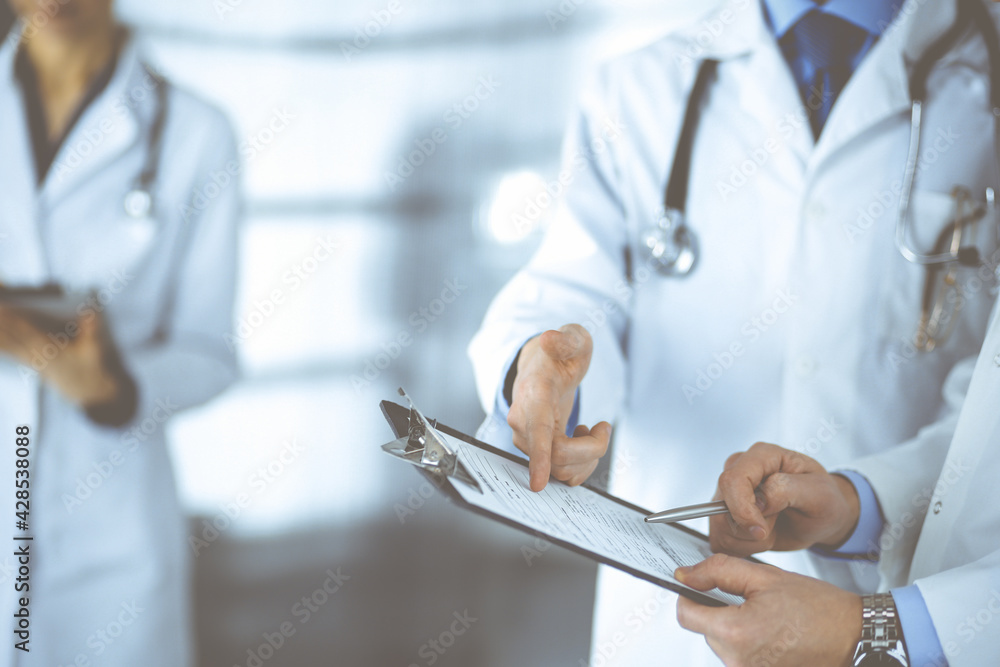 Unknown doctors, two men and a woman, discuss medical exam resoults, while standing at hospital office. Physicians using clipboards for filling up medication history records. Perfect medical service