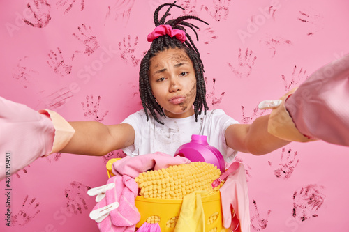 Unhappy overworked housewife stretches arms to make selfie stands dirty near basket full of laundry and detergents tired doing domestic duties isolated over pink wall. Housecleaning concept.