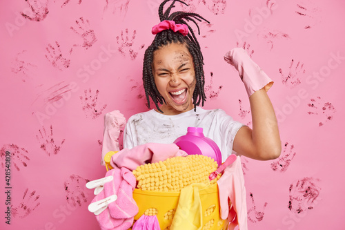 Happy housewife makes yes gesture clenches fists celebrates finishing house cleaning wears rubber gloves stands near laundry basket has fun isolated over dirty pink background. Yeah finally I am free