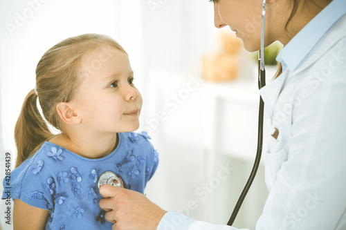 Doctor examining a child by stethoscope. Happy smiling girl patient dressed in blue dress is at usual medical inspection. Medicine concept