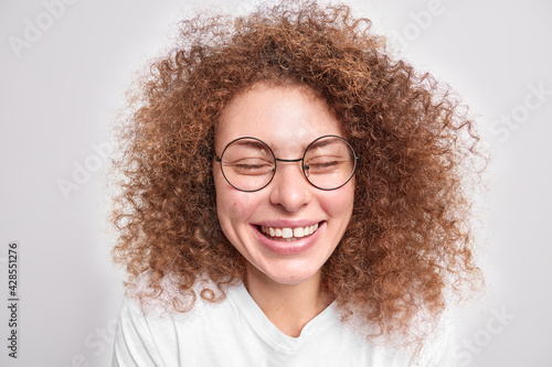 Close up portrait of lovely curly haired female model laughs happily keeps eyes shut wears round transparent glasses casual t shirt isolated over white background chuckles at camera stands carefree