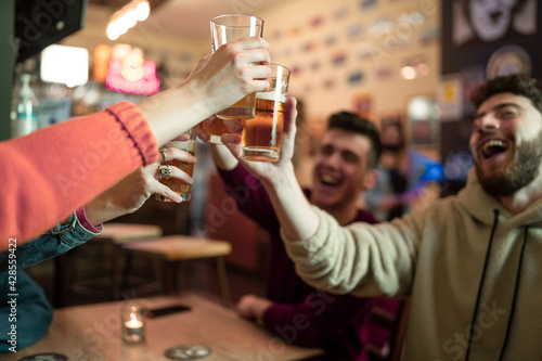Group of young people hangout in a pub restaurant having fun and toasting, clinking beer glasses. Youth culture of millennials toasting and drinking in the night time