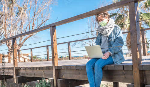 Spanish woman wearing a facemask with a laptop in her lap while sitting on a wooden bridge photo