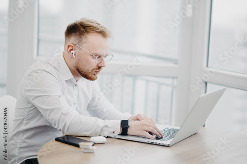 young man in glasses wearing wireless headphones works with laptop, video conference © velimir
