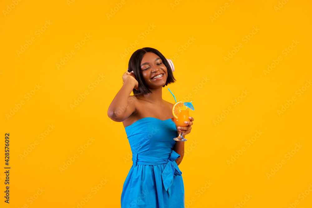 Black lady in casual outfit holding tropical summer cocktail and listening to music in headphones on orange background Black lady in casual outfit holding tropical summer cocktail and listening to music in headphones on orange background