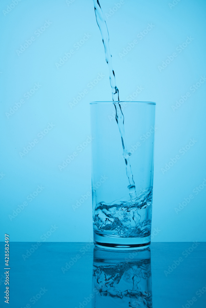 Glass of pure still water. Thirst quenching. Pure still water pours from bottle to glass against blue background with back lighting Glass of pure still water. Thirst quenching. Pure still water pours from bottle to glass against blue background with back lighting