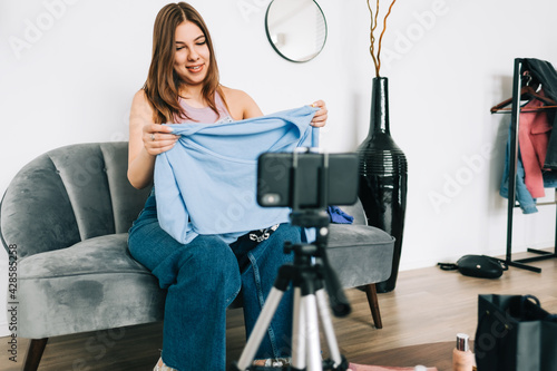 Attractive caucasian young woman video blogger showing new clothing via her blog on social media, holding blouse and looking at smartphone camera fixed on tripod.