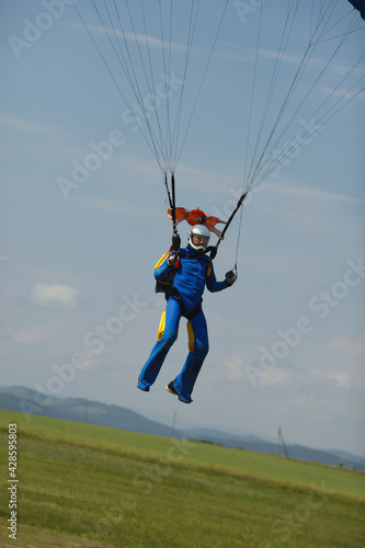 Skydiver figure under parachute slings during landing on a green field.