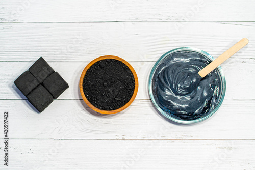 Charcoal, crushed fraction, homemade face mask and activated charcoal scrub of powder and yoghurt on white wooden background