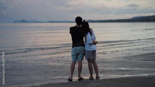 Back portrait of lovely couple stand on the beach and look at the horizontal line of seascape with sunset background
