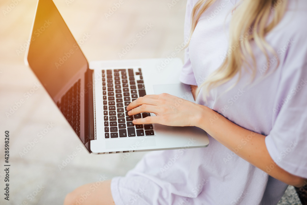 Fototapeta premium Young woman sitting on the city street typing message on laptop back view close-up. Woman hand on keyboard.