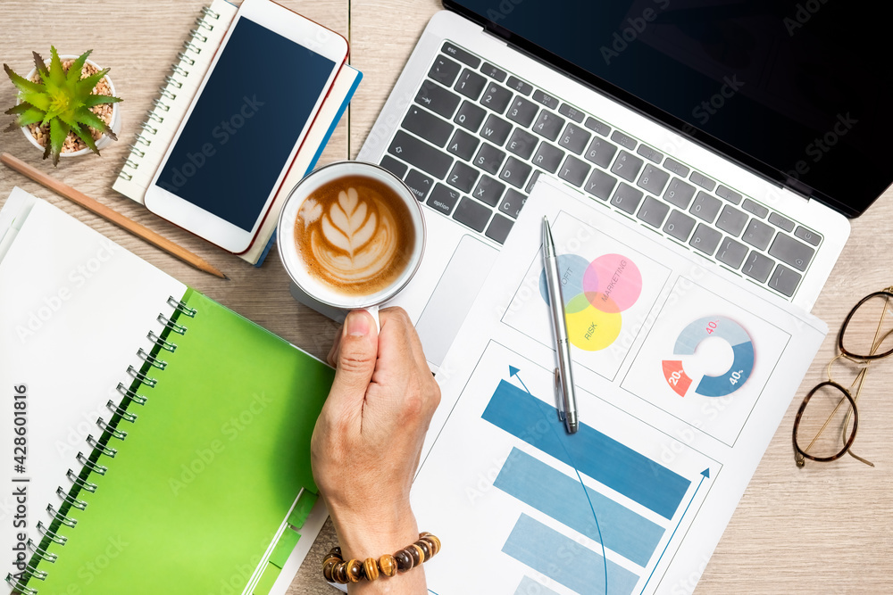 Man's hand holding coffee cup and smartphone, laptop and notebook on wooden table