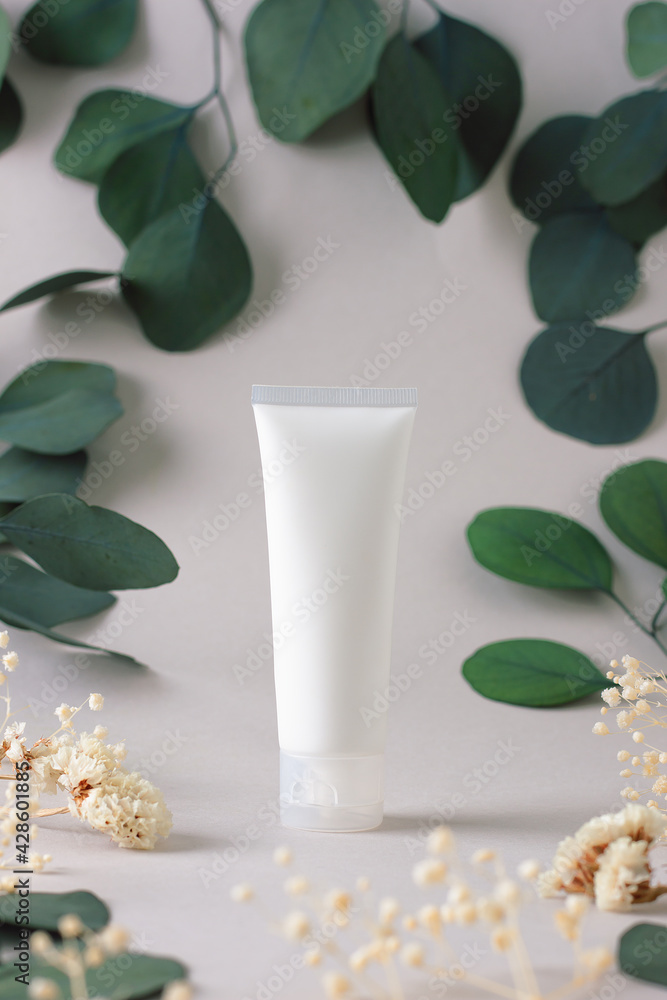 White tube of cosmetic cream with flowers and green leaves on grey background. Close up White tube of cosmetic cream with flowers and green leaves on grey background. Close up