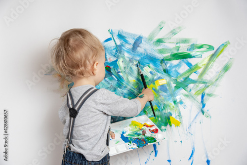 Little child with a brush and a palette of paints in his hands draws on a white wall. Happy childhood