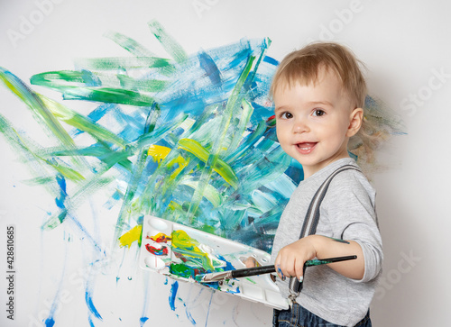 Little child with a brush and a palette of paints in his hands draws on a white wall. Happy childhood