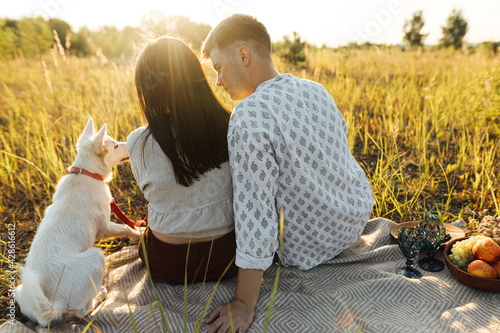Stylish couple with white dog relaxing on blanket in sunny light in summer meadow. Picnic with pet