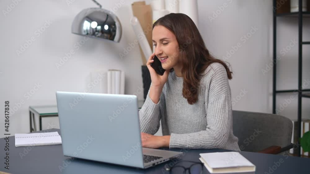 Positive young female entrepreneur in casual wear talking on the smartphone sitting at the desk, cheerful woman has phone conversation in unconstrained atmosphere in modern office space