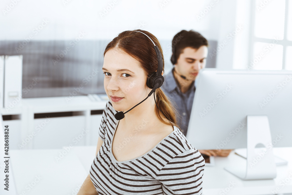 Casual dressed young woman using headset and computer while talking with customers online. Group of operators at work. Call center, business concept Casual dressed young woman using headset and computer while talking with customers online. Group of operators at work. Call center, business concept