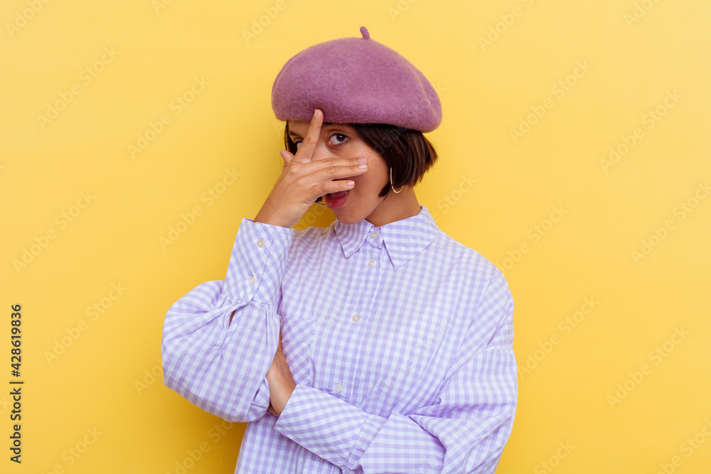 Young mixed race woman wearing a beret isolated on yellow background blink at the camera through fingers, embarrassed covering face. Young mixed race woman wearing a beret isolated on yellow background blink at the camera through fingers, embarrassed covering face.