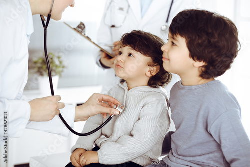 Woman-doctor examining a child patient by stethoscope. Cute arab toddler and his brother at physician appointment. Medicine concept