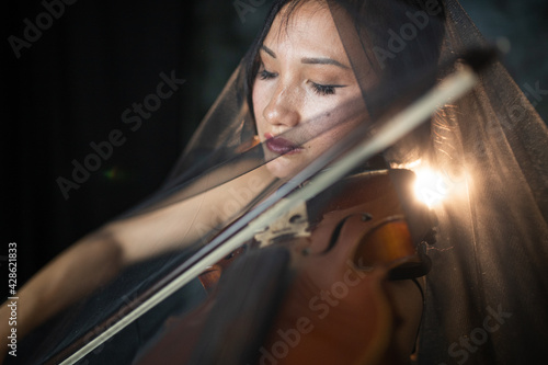 An Asian young woman is playing violin. Violinist is performing on the stage with the veil on her. Emotional playing on the musical instrument. Contour light. Artist.