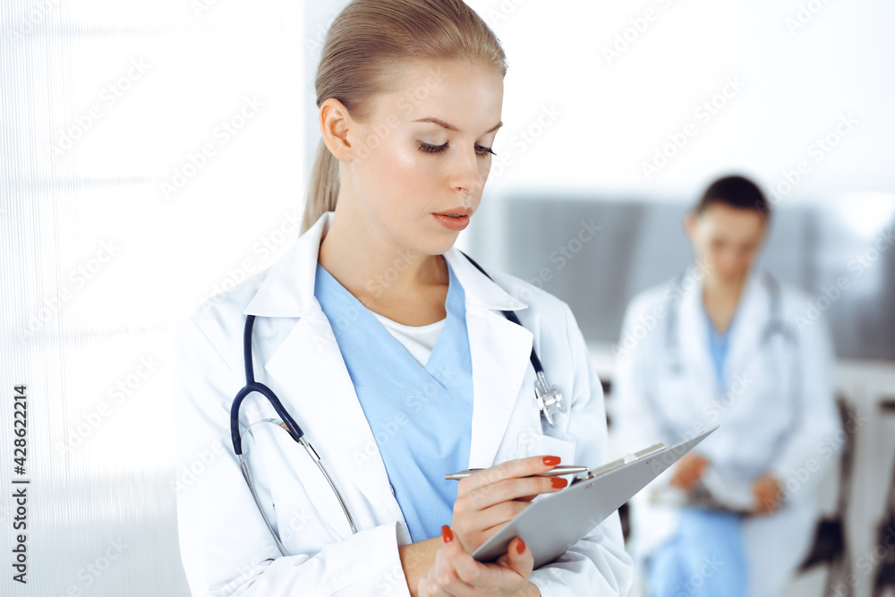 Woman - doctor is using clipboard for filling up medication history record in clinic with colleague at the background Woman - doctor is using clipboard for filling up medication history record in clinic with colleague at the background