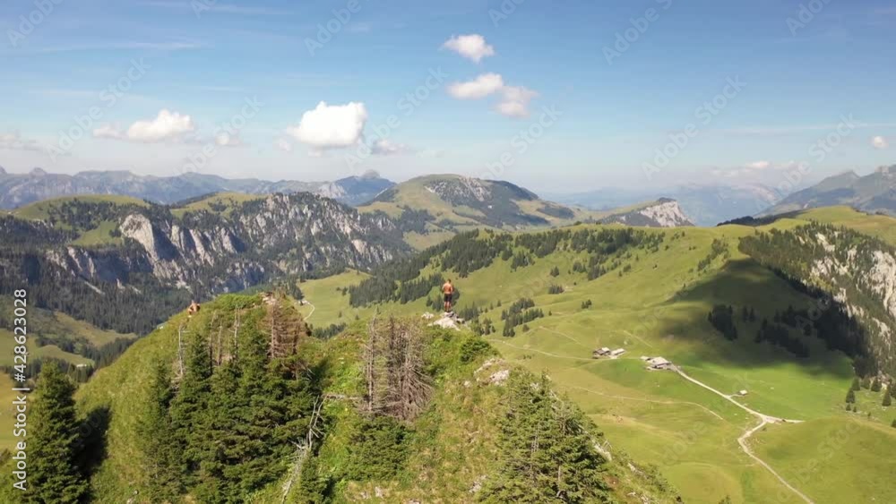 Drone circling around a young, strong and fit man standing on the edge overlooking a small, tiny lake below in beautiful turquoise and blue colors with green fields and forest around on a hot day. Drone circling around a young, strong and fit man standing on the edge overlooking a small, tiny lake below in beautiful turquoise and blue colors with green fields and forest around on a hot day.