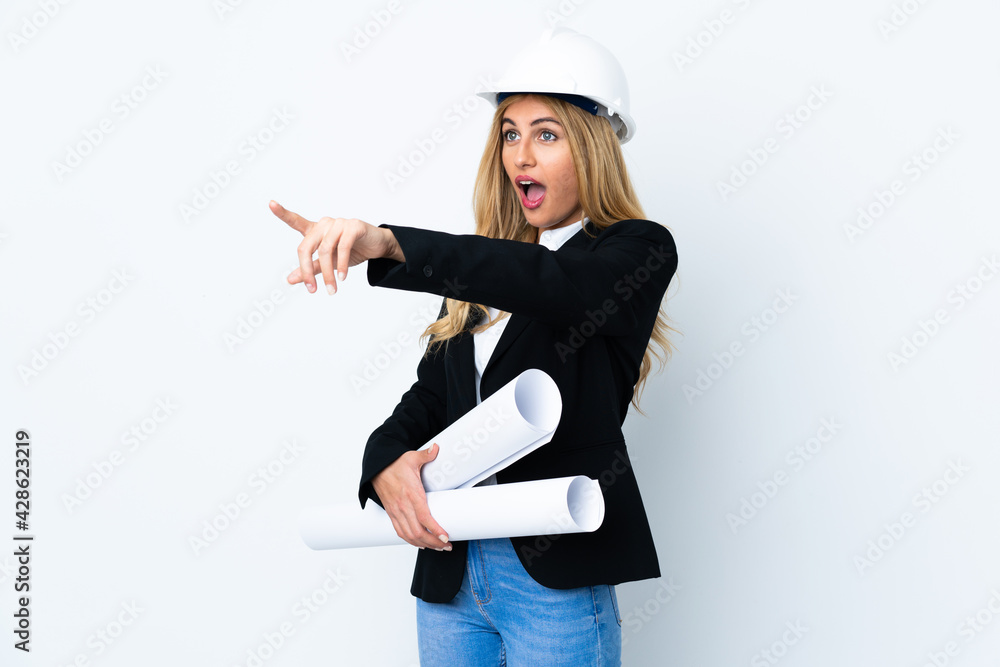 Young architect woman with helmet and holding blueprints over isolated white background pointing finger to the side Young architect woman with helmet and holding blueprints over isolated white background pointing finger to the side