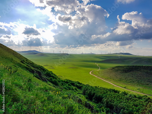 A panoramic view on a hilly landscape of Xilinhot in Inner Mongolia. Endless grassland with a few wind turbines between. Blue sky with thick, white clouds. A small gravel road through the pasture. photo