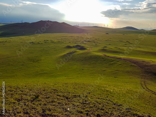 A panoramic view on a hilly landscape of Xilinhot in Inner Mongolia. Endless grassland with a few wildflowers between. The sun starts to set, coloring the sky orange. Blue sky with thick, white clouds photo