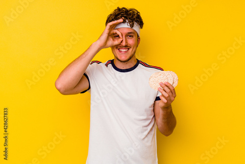 Young sport caucasian man eating a rice cake isolated on yellow background excited keeping ok gesture on eye.