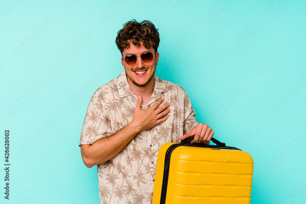 Young traveler man holding a yellow suitcase on blue background laughs out loudly keeping hand on chest.