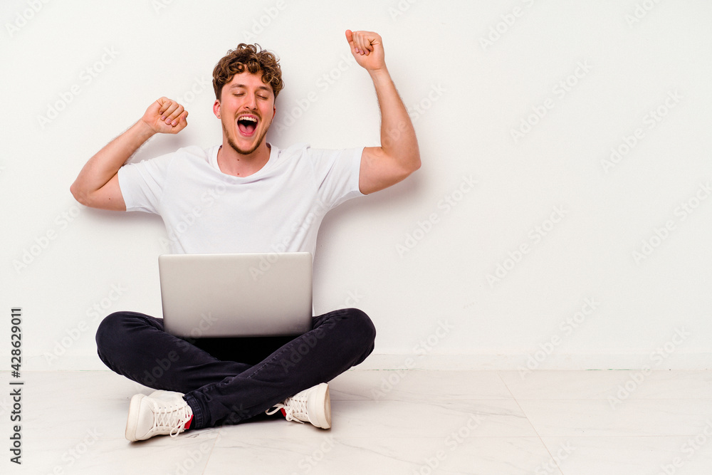Young caucasian man sitting on the floor holding on laptop isolated on white background celebrating a special day, jumps and raise arms with energy. Young caucasian man sitting on the floor holding on laptop isolated on white background celebrating a special day, jumps and raise arms with energy.